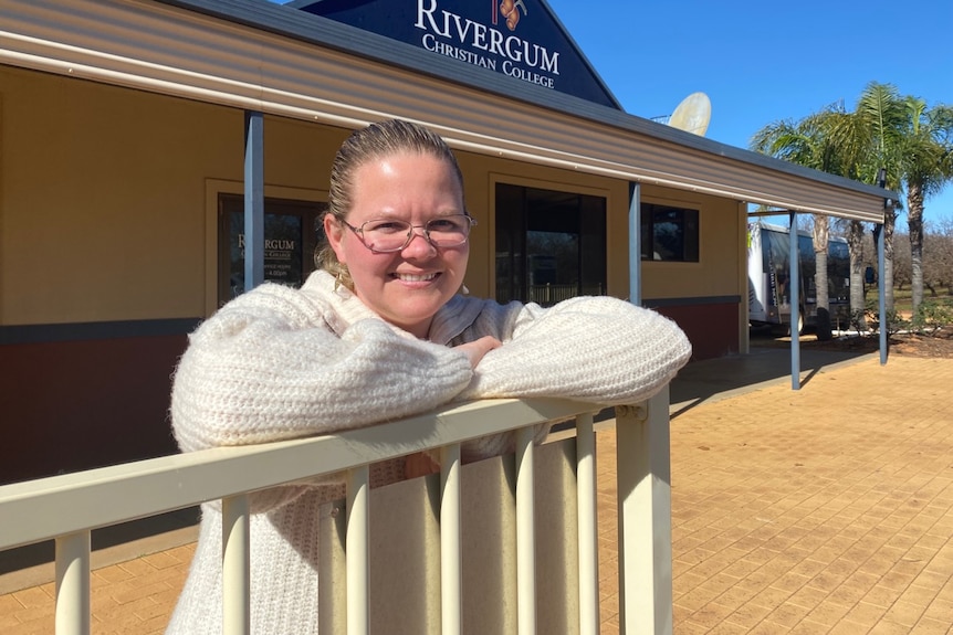 A woman wearing a white jumper leans against a fence in front of a sign that says Rivergum Christian College. 