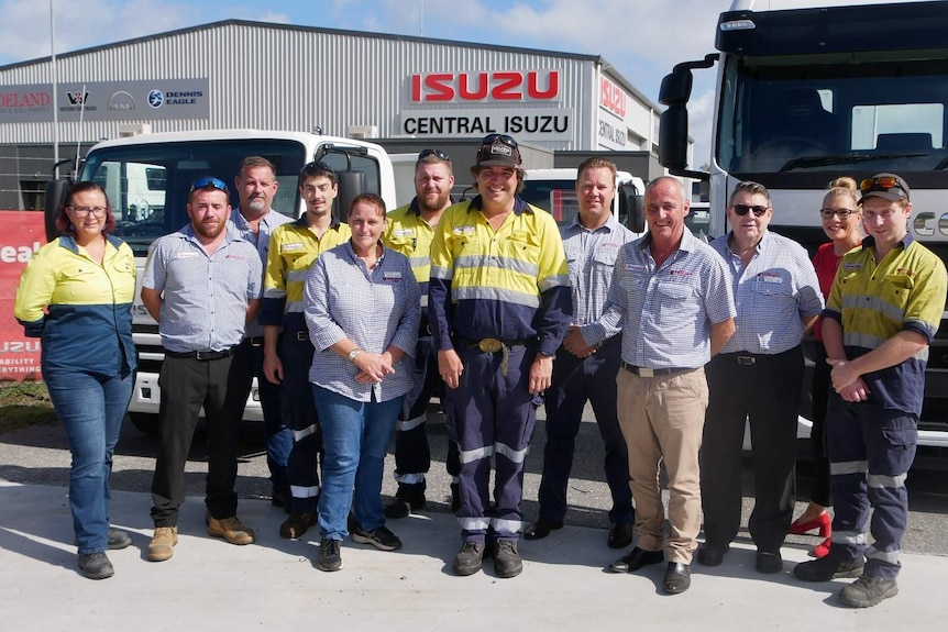 A group of 12 people, some in work overalls, smile as they stand in the sunshine with trucks and sheds in the background.