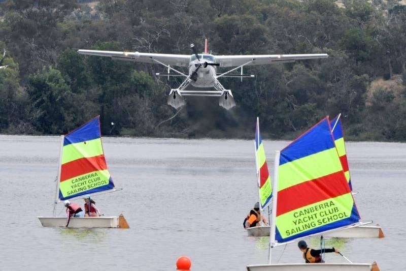 A seaplane lifts off the lake over the top of children in yachts.