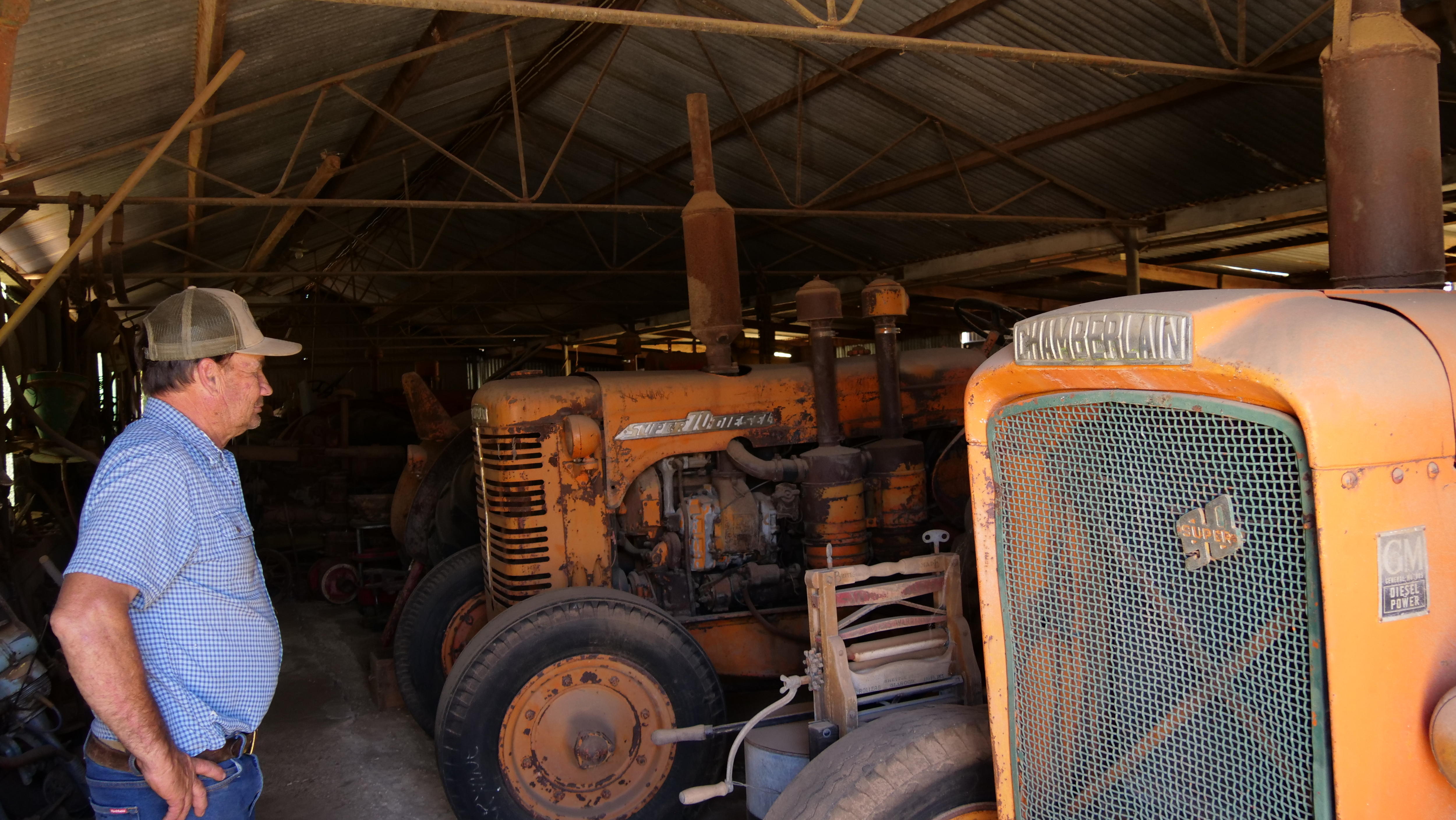 Um homem parado em um galpão, olhando para seu antigo maquinário agrícola. 