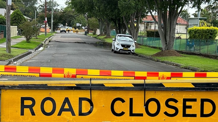 A road closed sign lays before a street with water pooled and a hole on one side.