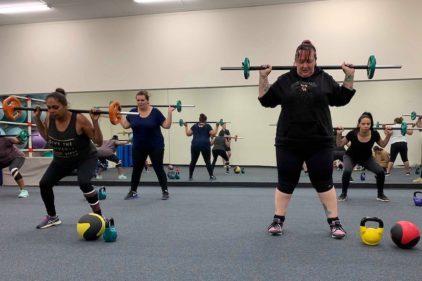 A group of women are lifting metal bars with weights above their heads. The weights are all different colours.