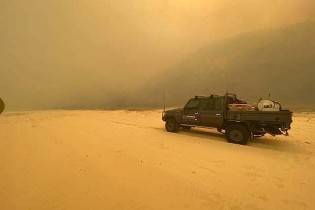 Vehicle drives along a beach, the sky is orange from wild fires burning 