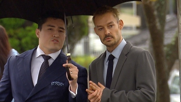 Two men stand under an umbrella waiting at traffic lights.
