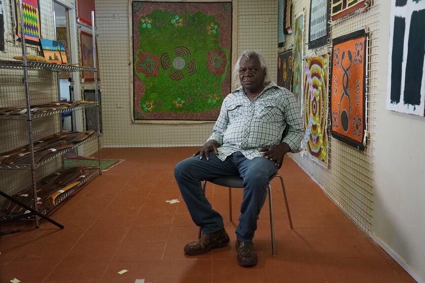 Peter Corbett sits on a chair in a room surrounded artworks on the walls.