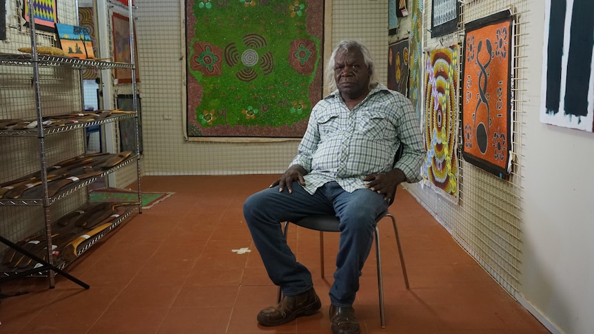 Peter Corbett sits on a chair in a room surrounded artworks on the walls.