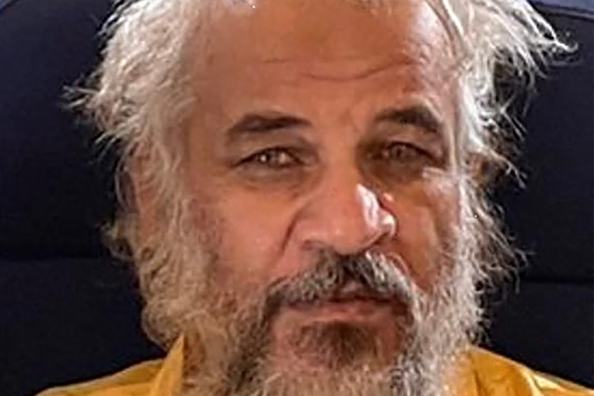 A bearded man with balding grey hair looks at the camera while sitting in a chair in a yellow shirt. 