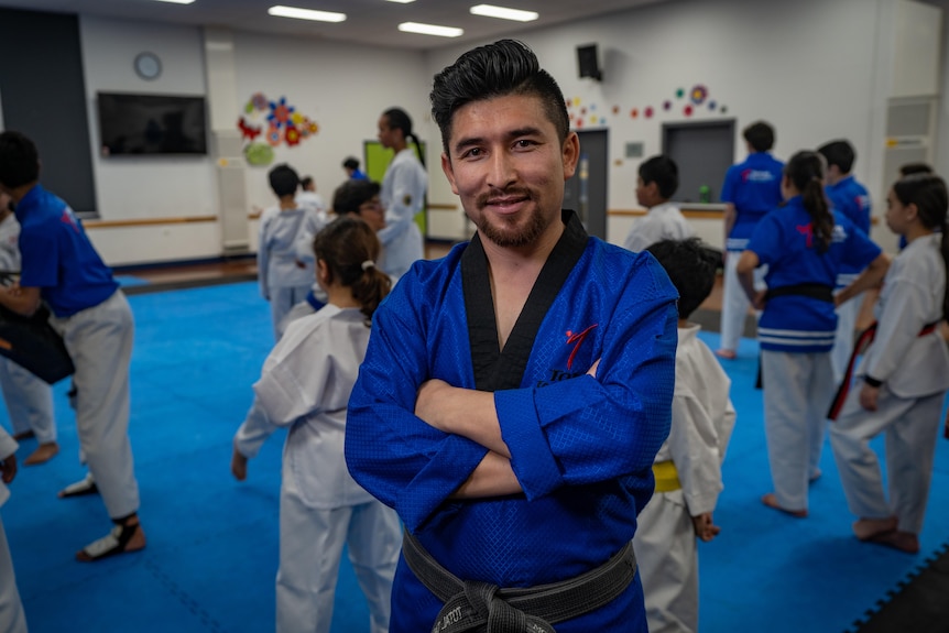 An Afghan man wearing a blue taekwondo outfit with slicked back hair smiles at the camera with children behind him