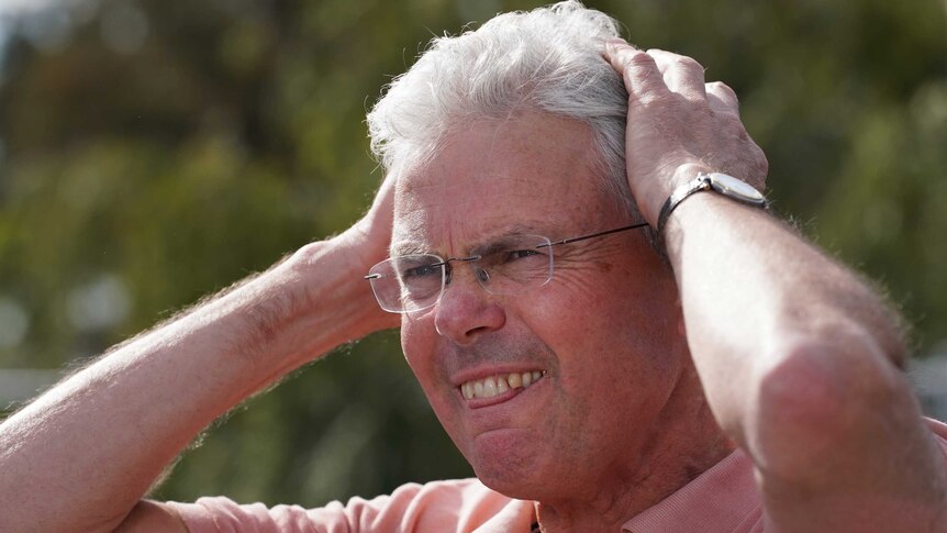 A racehorse trainer grimaces in disbelief as he puts both hands to his head in frustration.