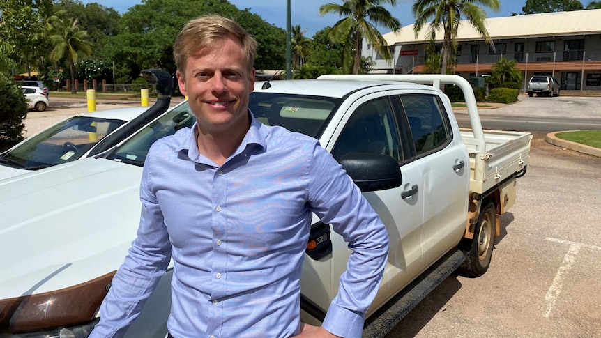 A blonde man with a long-sleeved blue shirt stands in front of a ute, with palm trees in the background