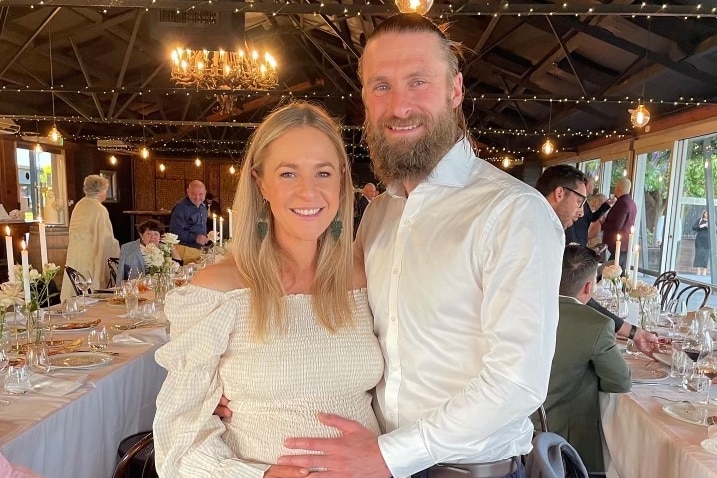 Young couple stands smiling at a wedding, male places hand on woman's pregnant belly.