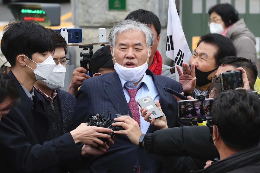 A man in a suit speaks over a lowered face mask to a group of surrounding media, who are all wearing face masks.