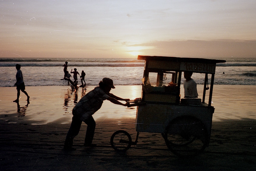 A street merchant wheels his cart along Kuta Beach in Bali while passersby look on.