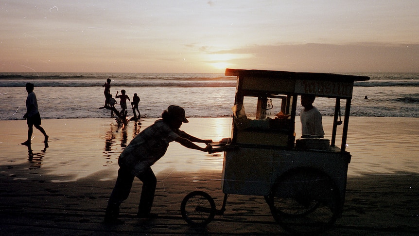 A street merchant wheels his cart along Kuta Beach in Bali while passersby look on.
