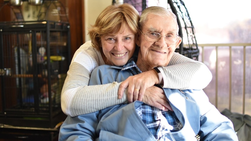 A woman wrapping her arms around her elderly father from behind while he is seated.