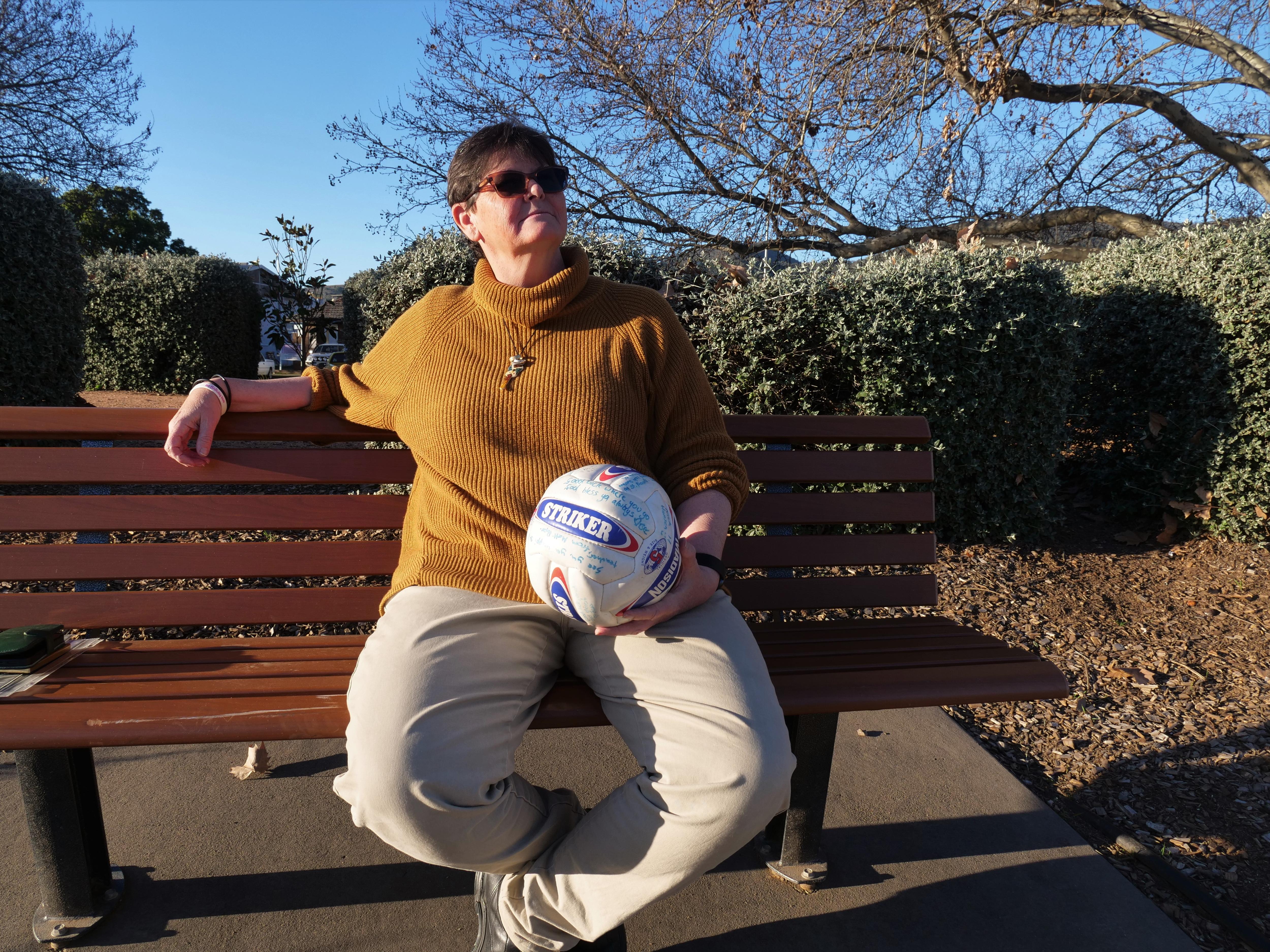 A woman with a turtle neck sweater enjoys the sun on a park bench with a signed soccer ball in her hands