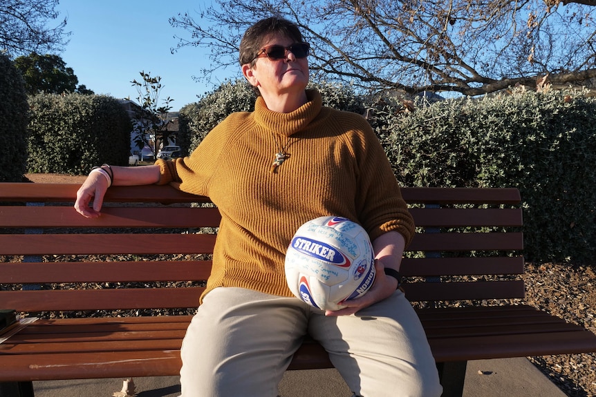 A woman with a turtle neck sweater enjoys the sun on a park bench with a signed soccer ball in her hands