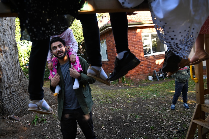 A man looks up at some children while holding a child on his shoulders in a suburban backyard.