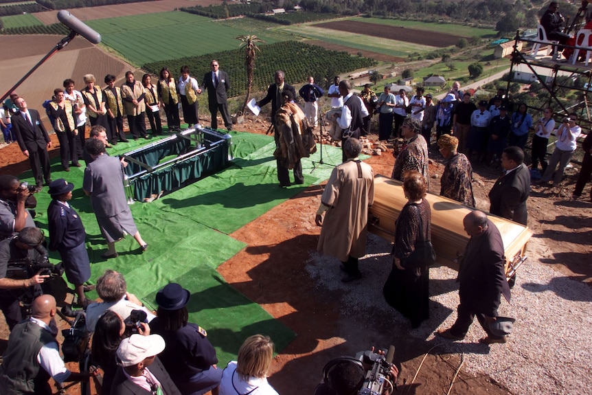 From above, a crowd is seen gathering near a coffin being carried and an open grave.