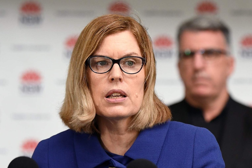 Kerry Chant, with shoulder-length blonde hair, spectacles and a dark top, addresses the media.