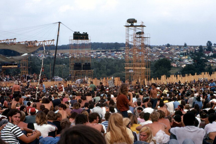 A sea of people sitting down, with a stage in the background and tents on a hill in the distance