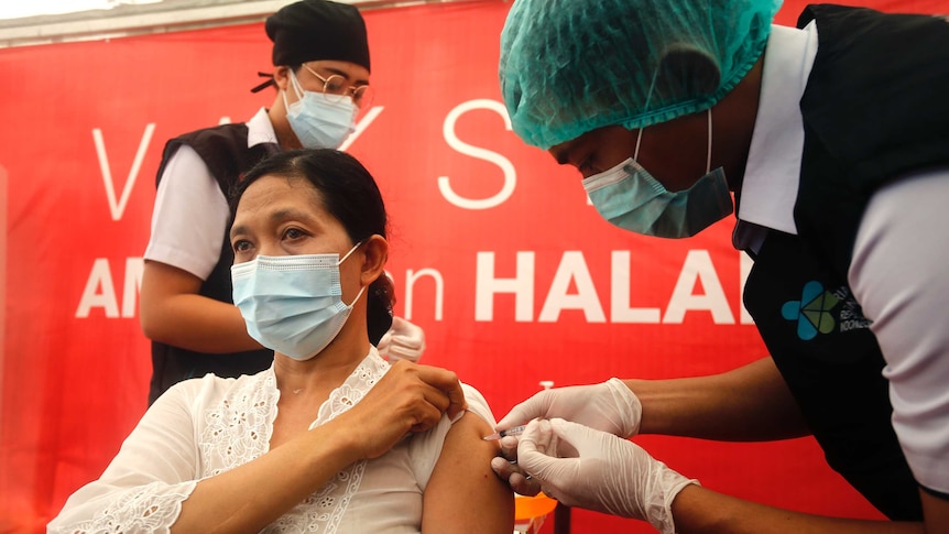 A woman sits in a chair wearing a mask as a man wearing a hairnet administers a needle of COVID-19 vaccine