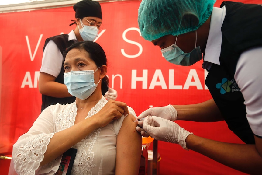 A woman sits in a chair wearing a mask as a man wearing a hairnet administers a needle of COVID-19 vaccine