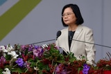 Taiwan's President Tsai Ing-wen stands behind a lecturn cpvered in flowers. Two microphones are pointed at her.