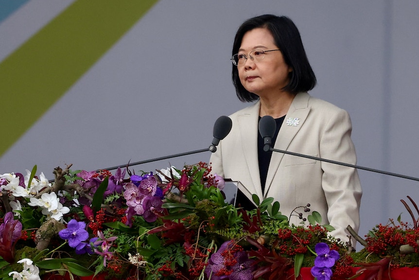 Taiwan's President Tsai Ing-wen stands behind a lecturn cpvered in flowers. Two microphones are pointed at her.