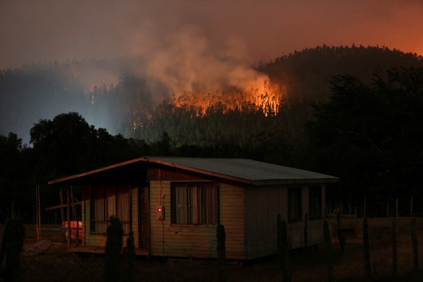 A fire seen behind a house in a mountain range with flames and smoke visible