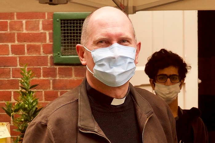 Father Hugh Kempster stands outside a brick building wearing a face mask and jacket.