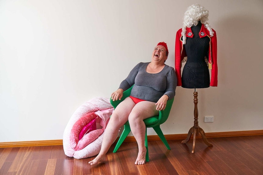 A woman in a grey T-shirt and red underpants sits in a chair, laughing