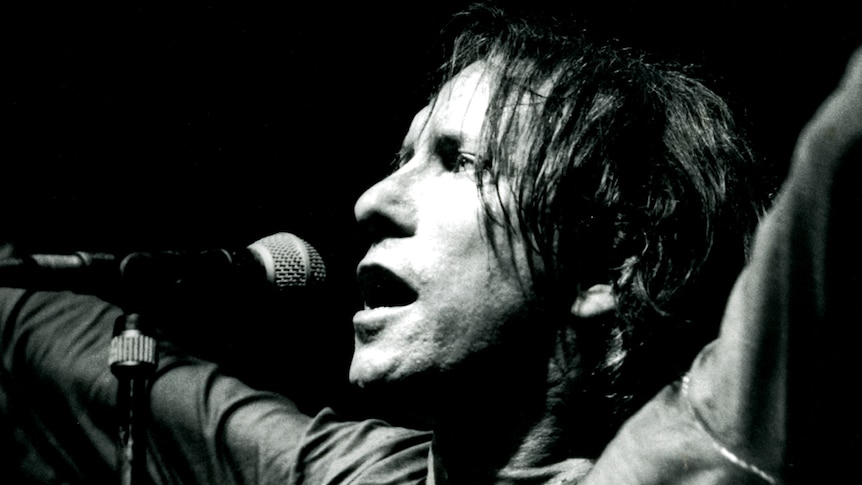 A close up black and white shot of Ron S. Peno standing in front of a microphone with his arms raised