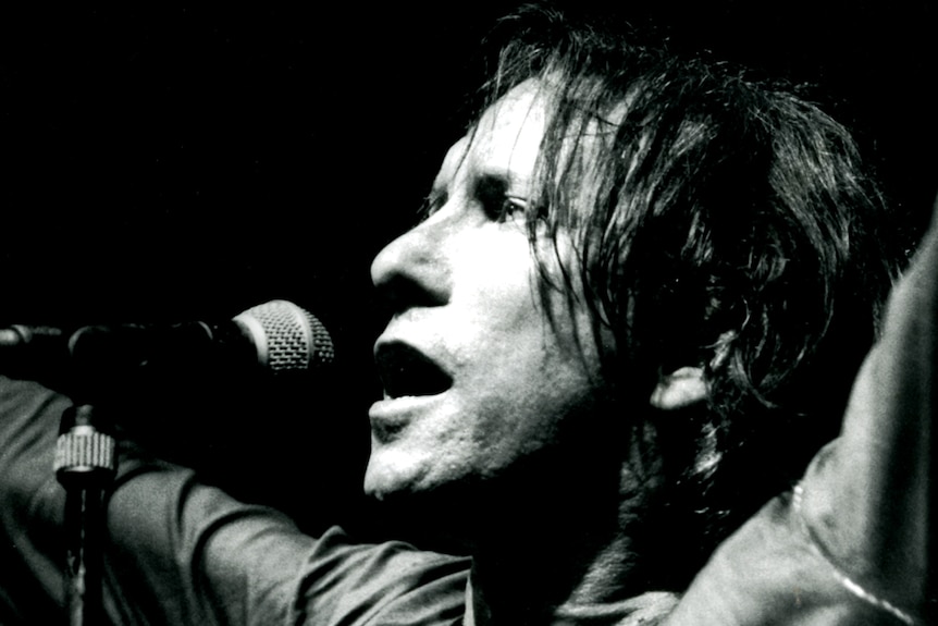 A close up black and white shot of Ron Peno standing in front of a microphone with his arms raised