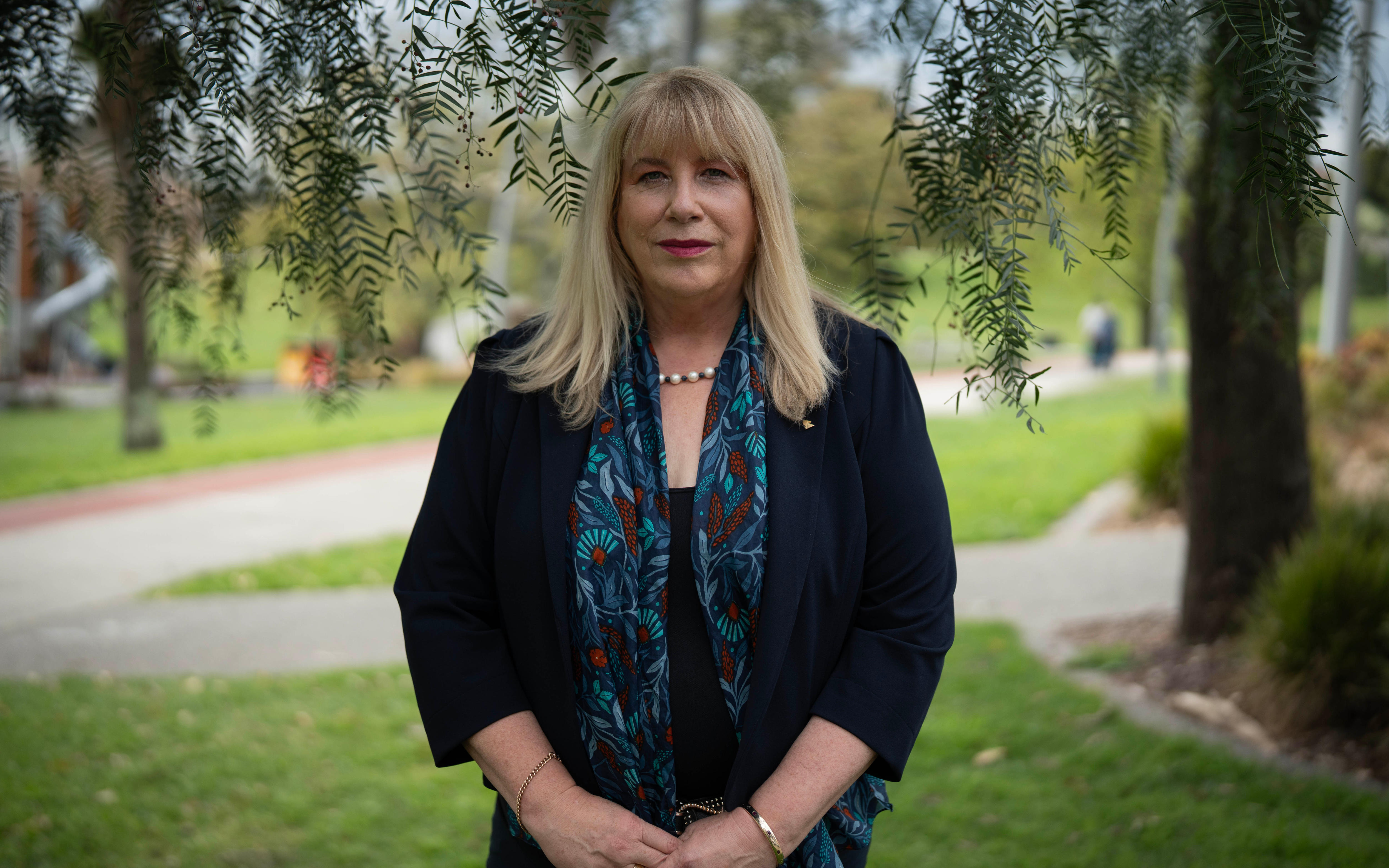 A woman with long blonde hair in a black top and blue patterned scarf stands in a park staring at the camera.