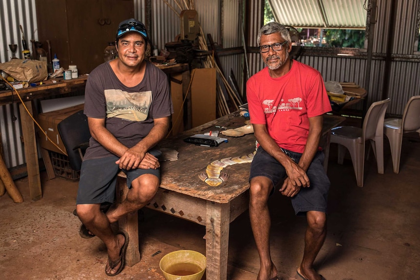 West Kimberley artists and brothers Garry and Darrell Sibosado sit on a low table in an art studio posing for a photo.