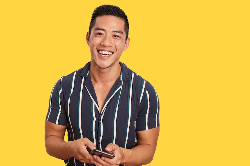 Photo of David Wang holding a phone on a yellow background, he's faced racism on gay dating apps like Grindr.