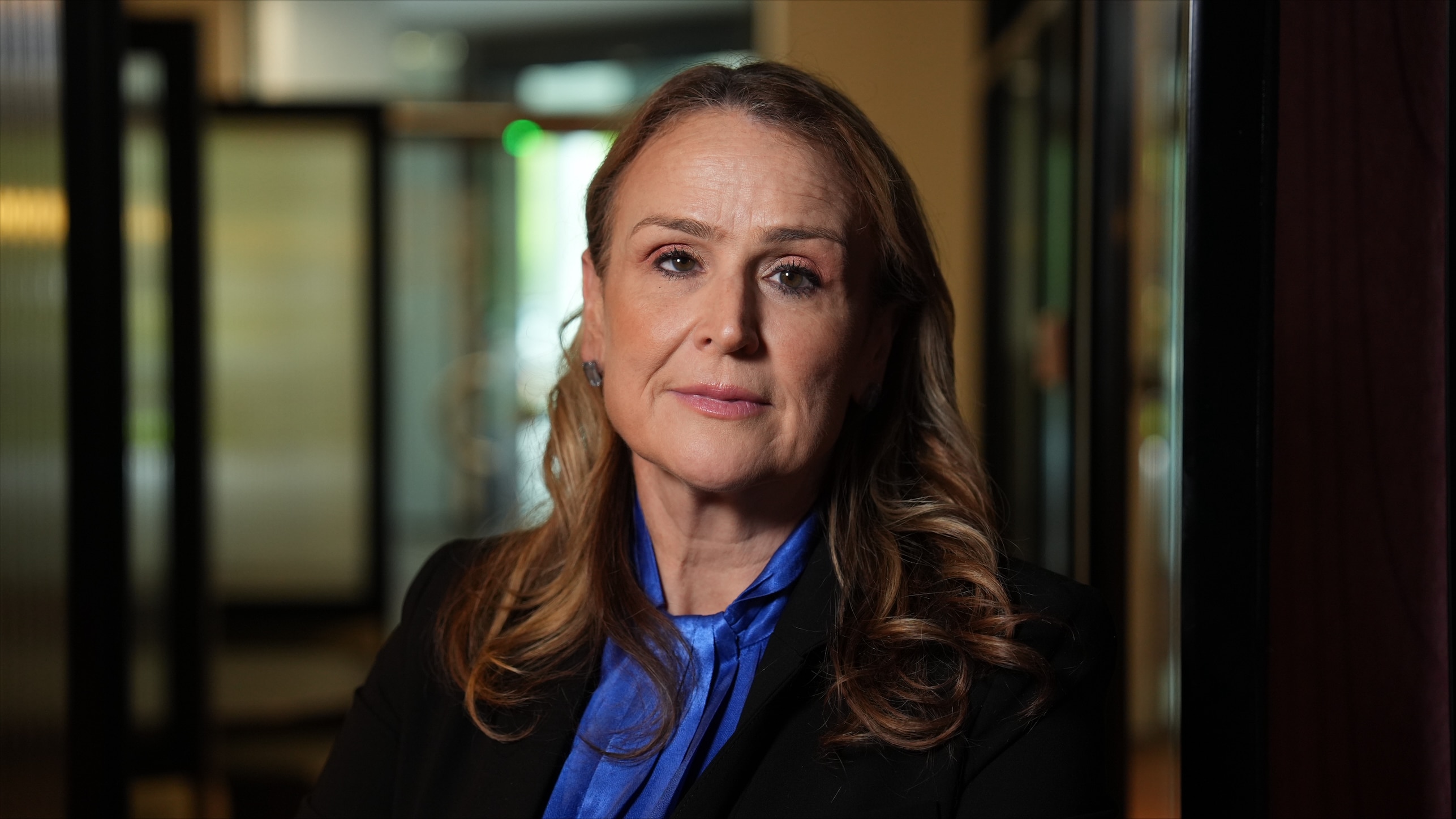 Misha Schubert, a woman with light brown hair, wears a black jacket over a blue shiny top with a serious facial expression.