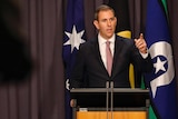 Chalmers raises a finger and points at media, an out of focus camera visible at the edge, standing in front of a blue curtain.