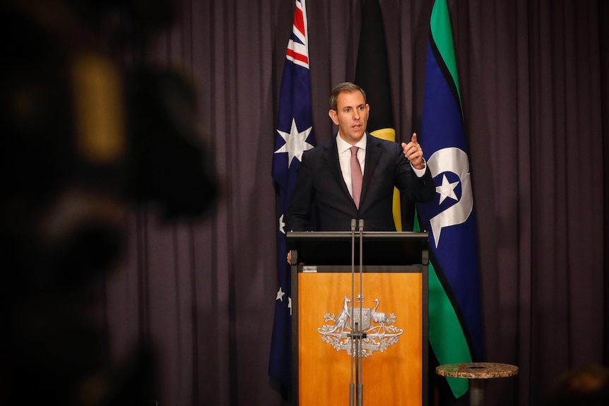 Chalmers raises a finger and points at media, an out of focus camera visible at the edge, standing in front of a blue curtain.