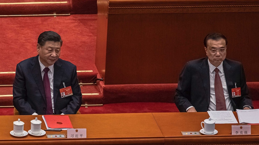 Two Asian men in suits sit behind desks in a chamber with red carpet.