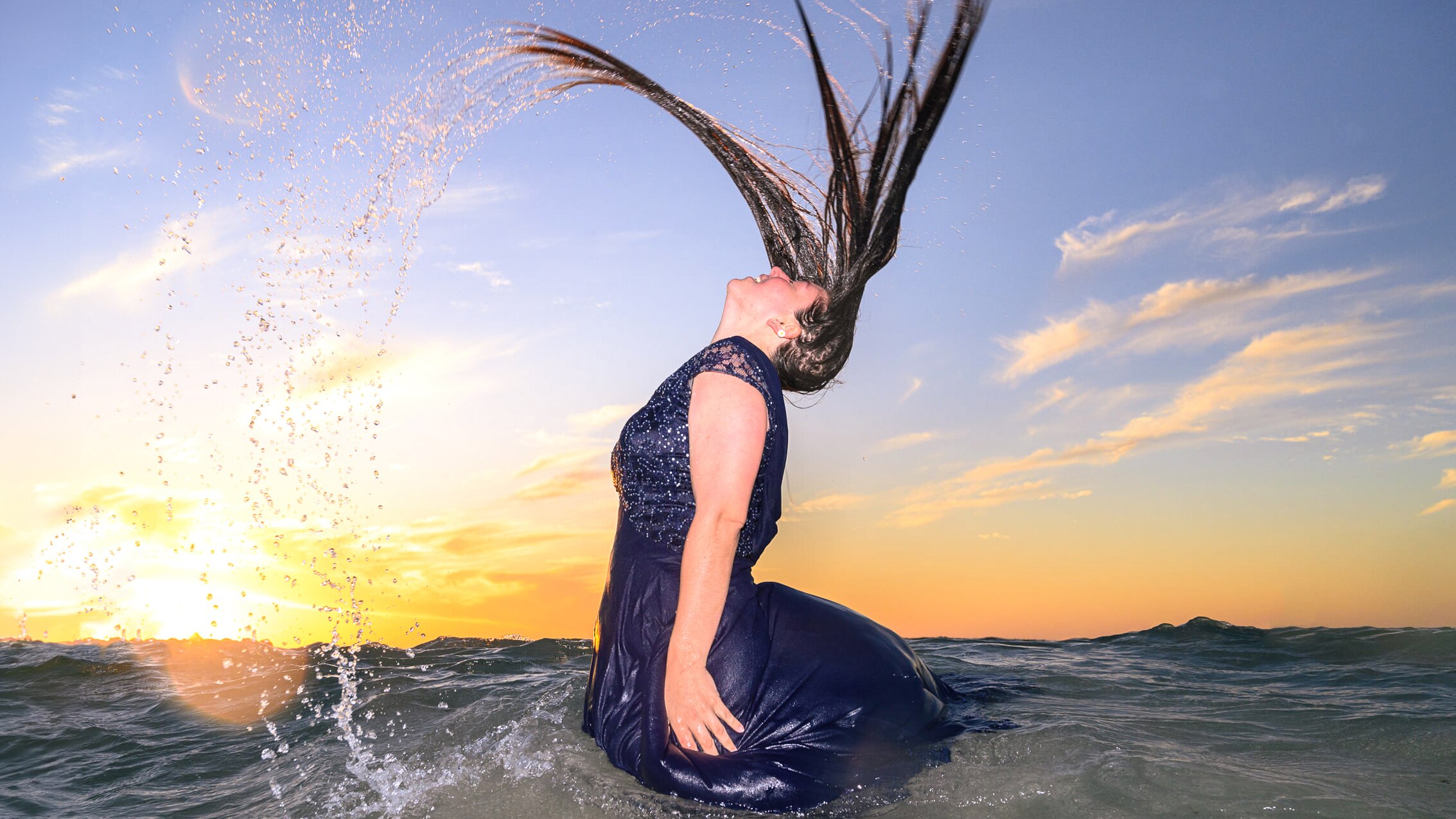 A woman in a navy dress flips her hair in the ocean.