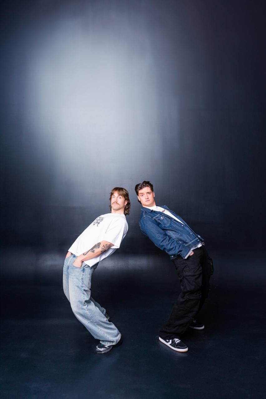 Two men wearing jeans and a tshirt lean back exageratedly into eachother 