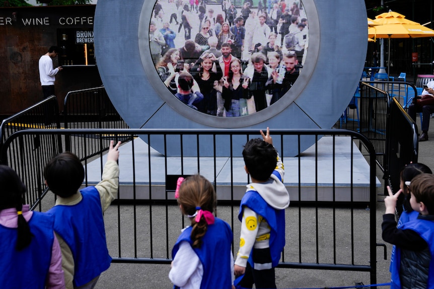 Kids are pictured in front of the portal making peace signs with their hands as people on the other side do the same.