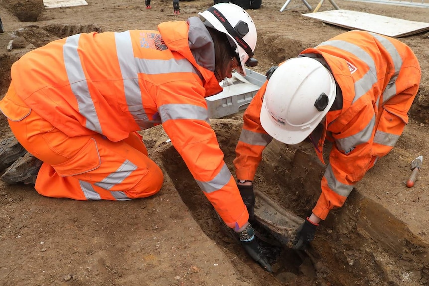 Two people in hard hats and high-vis work clothes lean over a dirt grave, carefully lifting out a breast plate