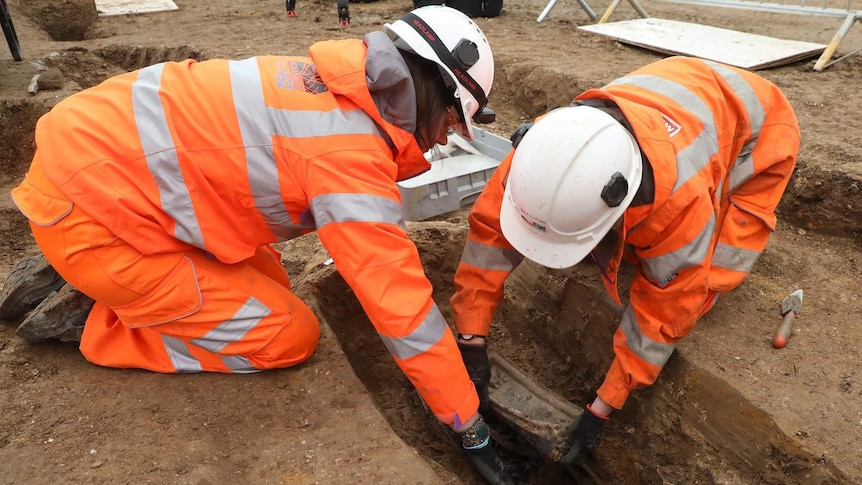Two people in hard hats and high-vis work clothes lean over a dirt grave, carefully lifting out a breast plate