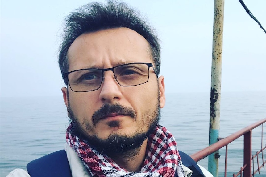 A man with glasses and checked scarf and goatee looking into distance with water in background.