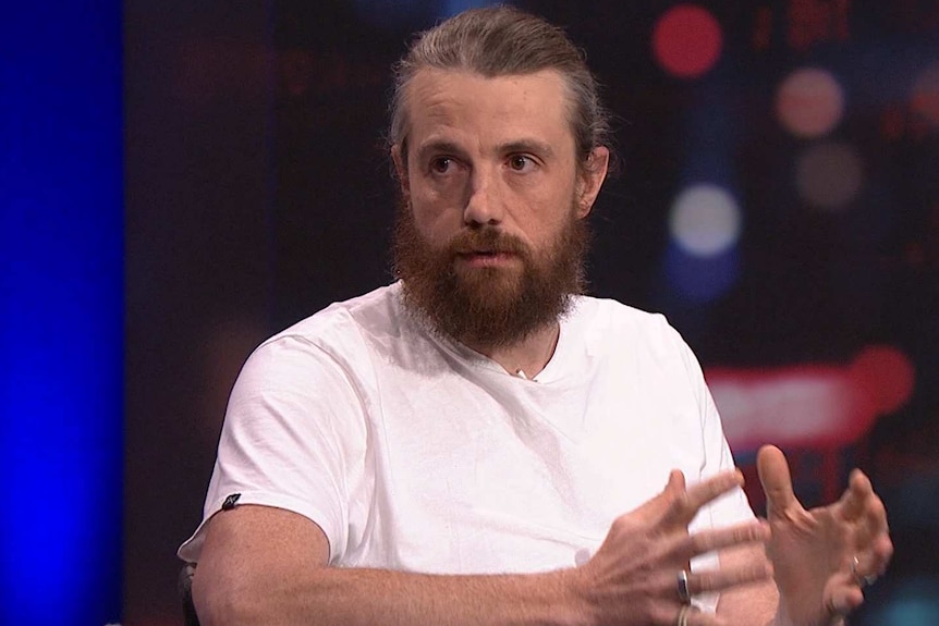 A man with a beard and his graying hair in a ponytail gesticulates with his hands, he is wearing a white t-shirt.