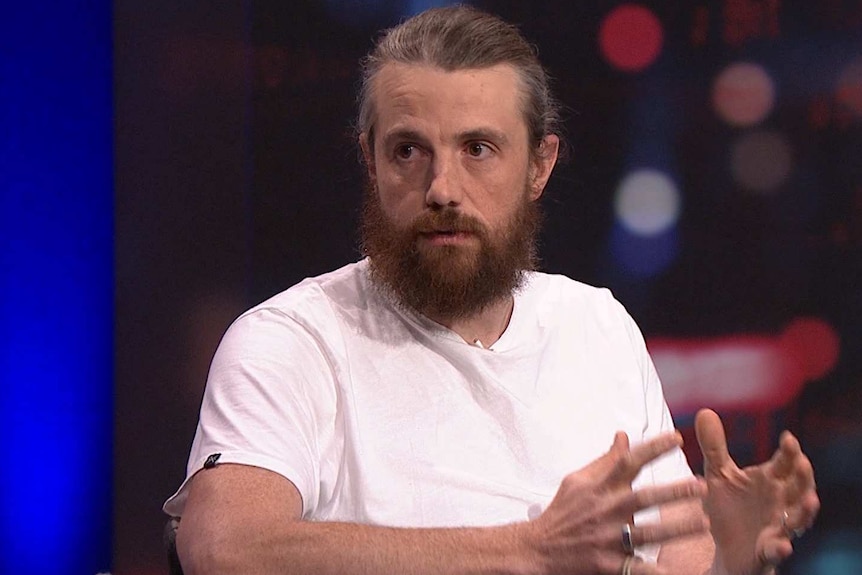 A man with a beard and his greying hair in a ponytail gesticulates with his hands, he is wearing a white t-shirt.