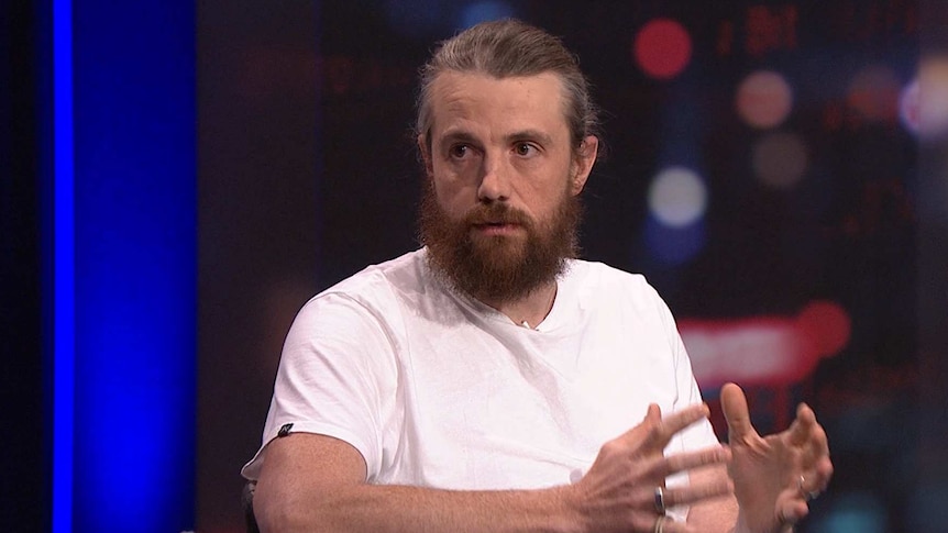 A man with a beard and his greying hair in a ponytail gesticulates with his hands, he is wearing a white t-shirt.